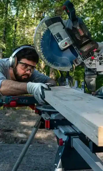 Wood artist Adam Detre stands surrounded by trees behind a Bosch GCM 18V-305 GDC Professional cordless crosscut and miter saw, carefully aligning a long piece of wood.