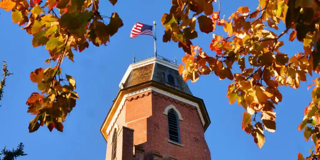 Brick tower with American flag, surrounded by vibrant autumn leaves, against a clear blue sky, conveying a peaceful, patriotic autumn scene.