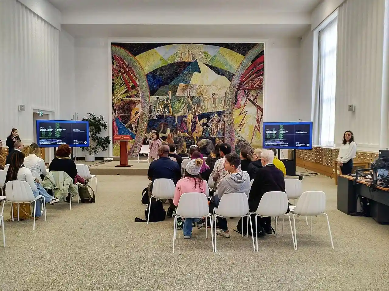 A group of Hack the Hackathon participants in the UN Libraries in Geneva. There's a huge, colourful painting in the background.