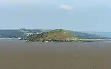 Green covered rocky land in expanse of sea. Hills behind.
