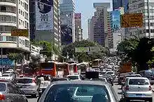Looking down a busy road, which is banked on both sides by tall buildings, some of which are covered in advertisement billboards