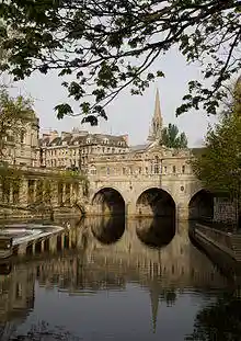 Yellow/Gray stone bridge with three arches over water which reflects the bridge and the church spire behind. A weir is on the left with other yellow stone buildings behind.