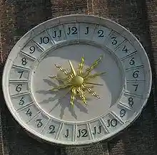 Decorative clay/stone circular off-white sundial with bright gold stylized sunburst in center of 24 hour clock face, one through twelve clockwise on right, and one through twelve again clockwise on left, with J shapes where ones' digits would be expected when numbering the clock hours. Shadow suggests 3 PM toward lower left.