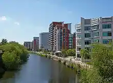 The canalised River Aire flows from the Dark Arches under Leeds's main railway station towards the bottom of the picture. To the left of the river is the lock which links the river with the Leeds and Liverpool Canal. To the right is a riverside walk beneath modern buildings, and in the distance, beyond the railway viaduct and station, are more high-rise modern buildings located on the west side of the city centre.