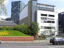 A quirky modern five-storey building with a large sign saying "BBC Yorkshire" in black above the second-floor windows on the white-fronted façade of the lower four floors can be seen on the far side of a dual-carriageway road with a barrier along the central reservation. At right-angles to the right of the building is a tall blue slab with the letters "BBC" in white at the top. The left side of the building is mostly brick-red with a few windows, but above it is a light blue windowless section. The roof above this and the grey fifth floor of the frontage curves gently down to the rear. A lone car is driving from left to right along the road; between it and the building, temporary boards have been erected in front of a building to the left. In the top left-hand corner of the picture, part of a tall many-windowed building can be seen.