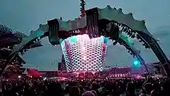 A concert stage; four large legs curve up above the stage and hold a video screen which is extended down to the band. The legs are lit up in green. The video screen has multi-coloured lights flashing on it. The audience surrounds the stage on all sides.