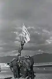 Israeli soldiers stabilize a flag pole whilst another soldier climbs it in order to raise an improvised flag; the soldier is seen about halfway up the flag pole. Other soldiers look on.