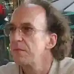 A middle-aged man with glasses and thinning, wavy brown hair looks to the left. He is wearing a light-colored shirt and appears to be outdoors, with blurred umbrellas and greenery in the background.