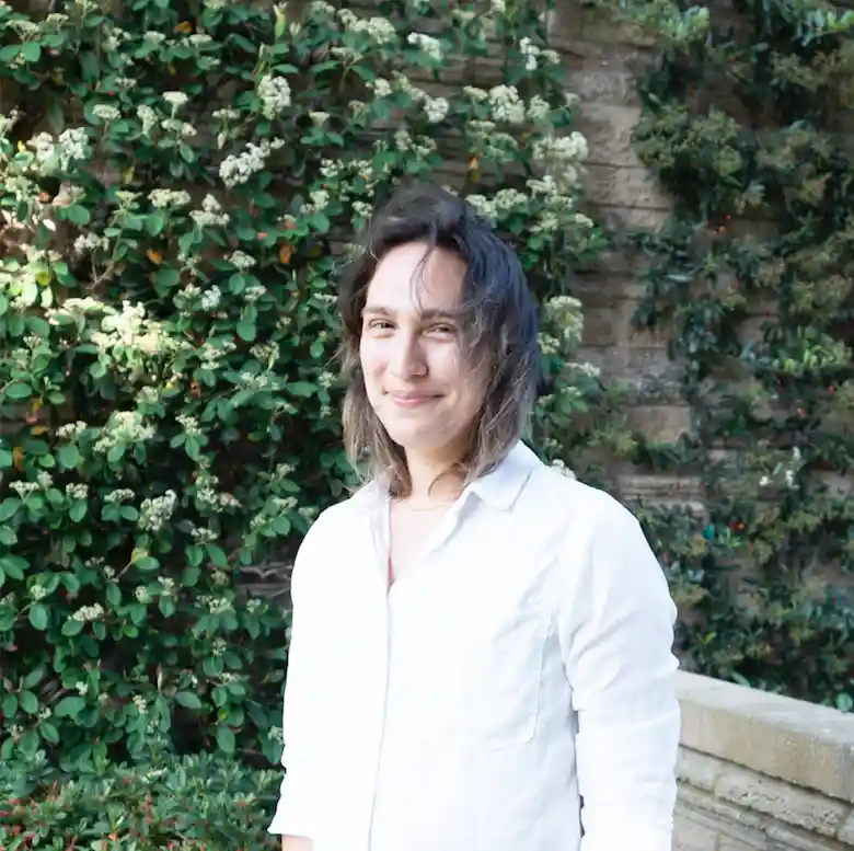 evelyn wearing a collared white shirt, standing in front of a stone wall covered in ivy. She is light skinned with shoulder length brown hair and looking at the camera, smiling slightly.