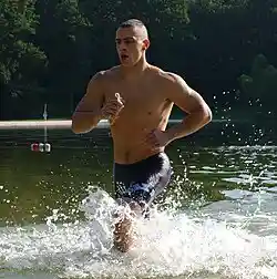 A photograph of a barechested man with short hair, running in shallow water wearing blue swimming trunks.