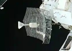 A rectangular dish shape of scaffolding covered in transparent sheeting, with a white insulation-covered radio receiver and support projecting from the centre. The blackness of space serves as the backdrop.