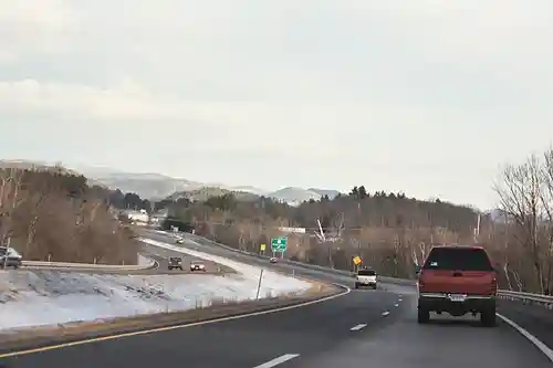 A four-lane highway in snowy weather, curving left with several cars on it. An exit sign and mountains are in the distance.
