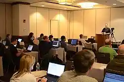 A man speaking from a podium to a room of people with laptop computers