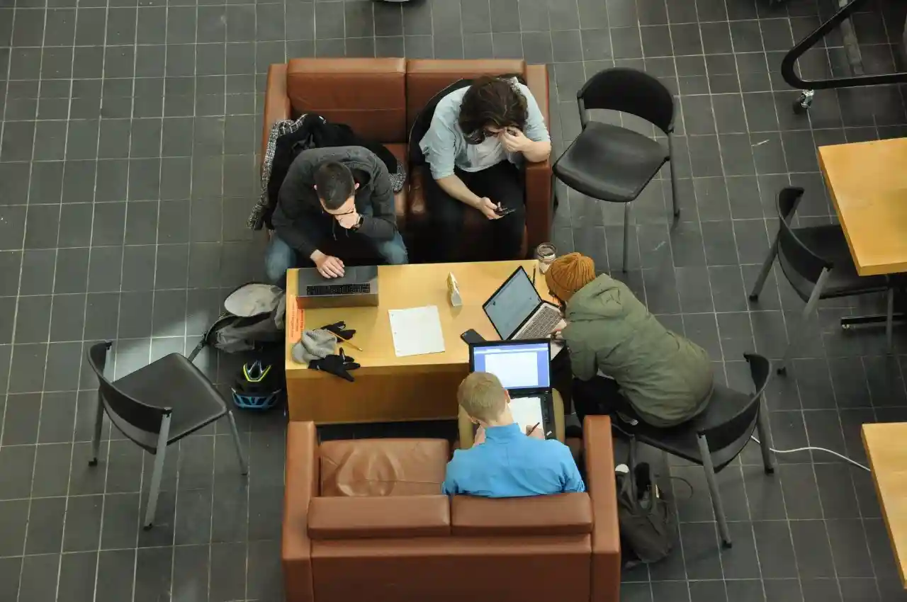 Studying in the bottom of the Computer Science building. Four students are shown from above sitting on chairs and sofas around a low table. Three are working on laptop computers and the fourth is looking at a mobile phone.
